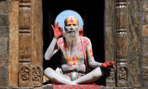 Homme assis en tailleur devant une arche sculptée, le corps peint de motifs colorés, la barbe longue et grise, levant une main en signe de bénédiction ou de mudra.