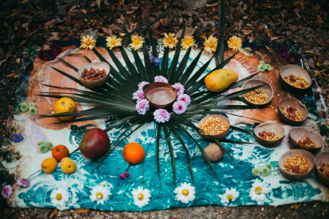 Installation rituelle en extérieur sur un tissu coloré, ornée de feuilles vertes en étoile, de fleurs jaunes et violettes, et de bols en bois remplis de grains de maïs et de fèves. Autour, des fruits variés comme mangues, oranges, abricots et banane sont disposés en cercle.