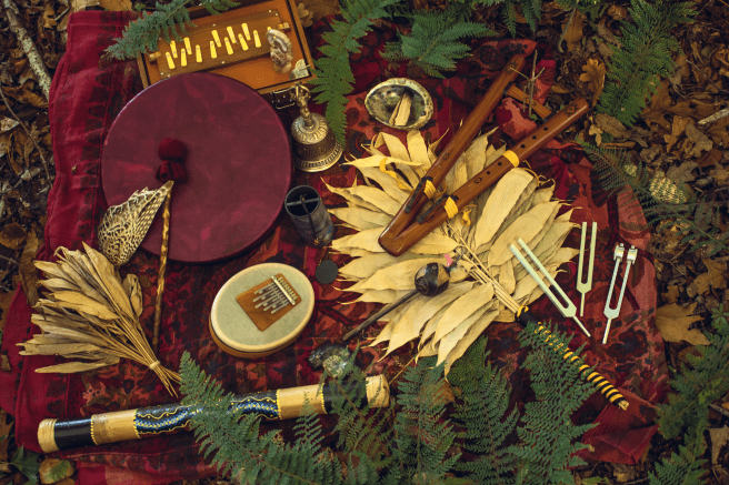 Ensemble d’instruments de thérapie sonore disposés en pleine nature sur un tissu rouge : tambour, flûte en bois, diapasons, kalimba et objets naturels, illustrant une pratique de bien-être, d’accompagnement thérapeutique et d’harmonisation corps-esprit