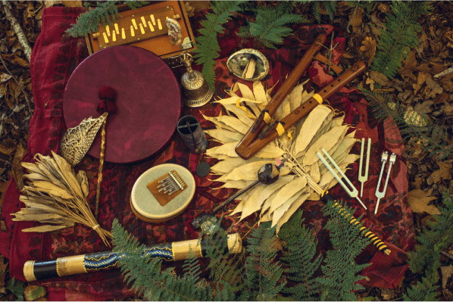 Ensemble d’instruments de thérapie sonore disposés en pleine nature sur un tissu rouge : tambour, flûte en bois, diapasons, kalimba et objets naturels, illustrant une pratique de bien-être, d’accompagnement thérapeutique et d’harmonisation corps-esprit
