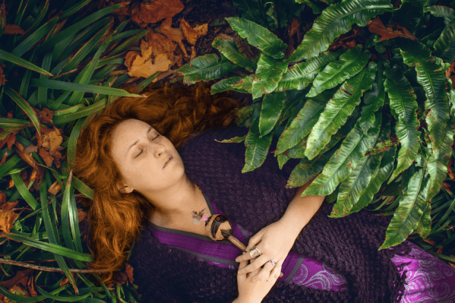 Sarah Stulzaft Vitalenergy, femme aux cheveux roux, vêtue d’une robe violette, est allongée sur le sol couvert de feuilles mortes et de plantes vertes. Elle semble dormir, les mains croisées sur sa poitrine en tenant un petit objet en bois orné de plumes.
