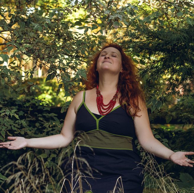 Sarah Stulzaft Vitalenergy, femme aux cheveux roux, debout en plein air dans une forêt, les bras ouverts et le visage levé, semblant profiter de la nature autour d’elle.