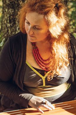 Femme aux cheveux roux, assise à l’extérieur, portant un collier de perles rouges et un gilet foncé, regardant vers le bas en posant une main sur une surface en bois, dans une lumière chaude et naturelle.