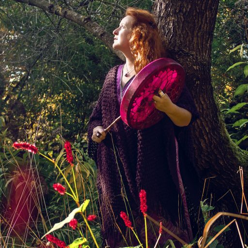 Sarah Stulzaft Vitalenergy, femme rousse debout dans une forêt, appuyée contre un arbre, regardant vers le ciel tout en tenant un tambour rose et une baguette ; elle porte une longue tenue sombre et est entourée de plantes et fleurs rouges.