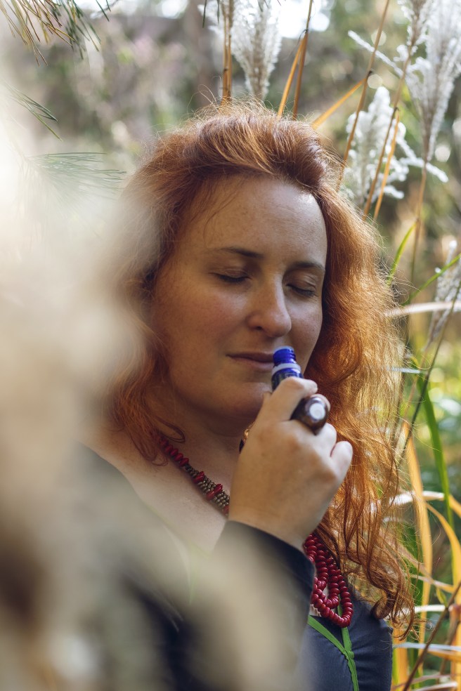 Sarah Stulzaft Vitalenergy, femme aux cheveux roux, les yeux fermés, respirant le parfum d’un petit flacon d'huiles essentielles dans un cadre naturel, entourée de végétation.